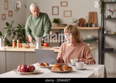 Femme âgée assise à la table avec un ordinateur portable et travaillant en ligne avec son mari cuisiner en arrière-plan dans le cuisine Banque D'Images