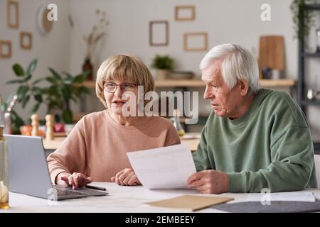 Couple senior assis à la table et utilisant un ordinateur portable pour payez vos factures en ligne dans la cuisine Banque D'Images