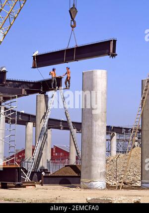 Historique 1984 archive view des travaux en cours sur le chantier de construction du bâtiment du projet de transport d'infrastructure de génie civil bâtiment des années 1980, Canary Wharf section aérienne du chemin de fer London Docklands Light LDR une grue abaissant des poutres en acier sur des colonnes de béton avec deux hommes travaillant sur des échelles sans règles modernes de santé et de sécurité une image d'archive de la façon dont nous étions dans l'est de Londres Angleterre au Royaume-Uni dans les années 80 Banque D'Images