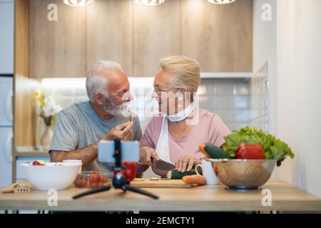 Couple senior enregistrant la vidéo sur la nourriture végétarienne sur la caméra dans la cuisine.Blogging concept. Les gens, la nourriture et les boissons, la technologie et le concept de style de vie Banque D'Images