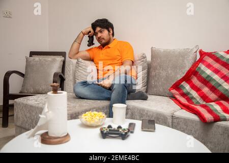 Homme indien asiatique jouant, diffusant et regardant la télévision seul dans le salon sur un canapé gris et une table blanche. Banque D'Images