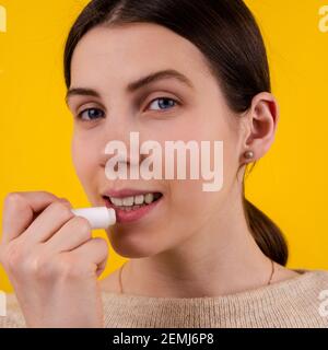 Belle jeune femme souriante utilisant un rouge à lèvres hygiénique sur fond jaune. Soin et protection des lèvres. Femme appliquant le baume sur les lèvres Banque D'Images