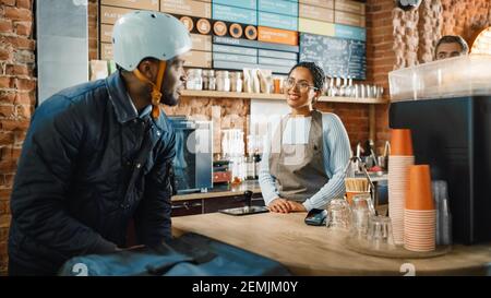 Courrier de livraison de nourriture afro-américaine parle à un jeune barista dans le restaurant moderne Bright Cafe. Le personnel était heureux et varié Banque D'Images