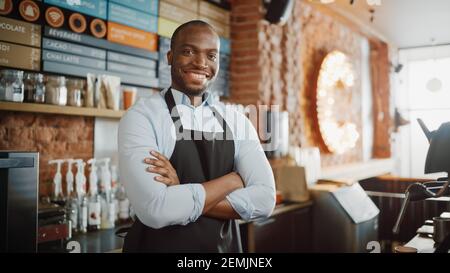 Le beau Barista afro-américain aux cheveux courts et à la barbe en tablier est souriant au café-restaurant. Portrait d'un employé heureux Banque D'Images