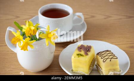 Tasse de thé et dessert de Pâques - petit farines de massepain jaune citron sur une assiette en porcelaine blanche pour le petit déjeuner et jonquilles sur une table en bois. Gros plan, Banque D'Images