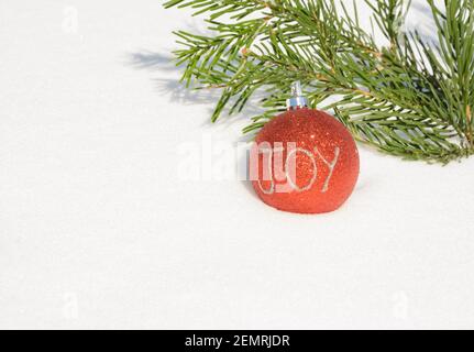 Boule rouge sur la neige fraîche avec une brindille de l'arbre de Noël, étincelante en plein soleil, avec TEXTE DE JOIE écrit sur la boule; avec espace de copie Banque D'Images