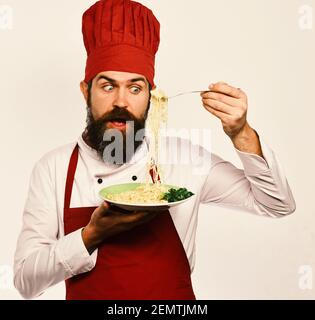 Concept de restauration rapide. Le chef mange des nouilles italiennes ou asiatiques. L'homme avec la barbe tient un plat savoureux sur fond blanc. Cuire avec le visage surprise dans l'uniforme bordeaux tient la fourchette et l'assiette. Banque D'Images