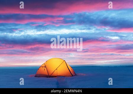 Tente jaune éclairée de l'intérieur par une lampe de poche sur fond de ciel rose gladeux. Paysage enneigé incroyable. Camp de touristes en hiver. Concept de voyage Banque D'Images