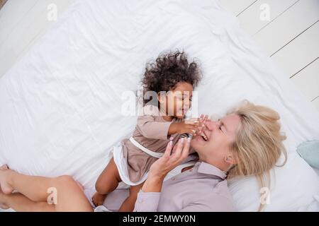 La jeune mère blonde caucasienne tickle sa petite fille afro-américaine. Allongez-vous sur un lit blanc, en vous amusant, avec vue sur le dessus. Les filles rient, embrassent, la femme kis Banque D'Images