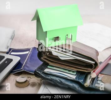 Au-dessus d'une pile de portefeuilles se trouve une petite maison en papier vert. Sur la table, tout autour, pièces de monnaie, divers papiers, crayon et stylo Banque D'Images