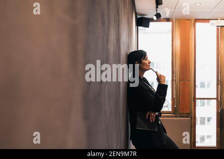 Femme entrepreneure mûre et réfléchie penchée sur le mur au bureau Banque D'Images