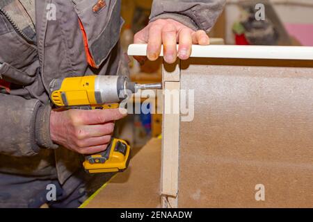 Le travailleur en charpenterie utilise un outil électrique manuel professionnel, un foret acu, et tord les vis sur le cadre de porte en bois. Banque D'Images