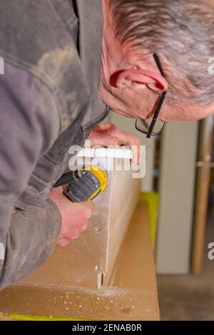 Le travailleur en charpenterie utilise un outil électrique manuel professionnel, un foret acu, et tord les vis sur le cadre de porte en bois. Banque D'Images
