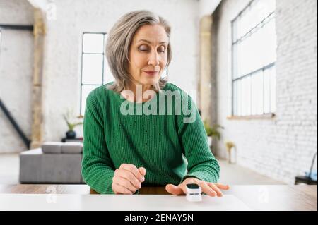 Femme âgée en bonne santé avec des rides sur le visage, vêtu de pull vert chaud, détermine la saturation des poumons avec de l'oxygène, à la maison, appartement de style loft, sur l'isolement, se soucier de sa santé Banque D'Images