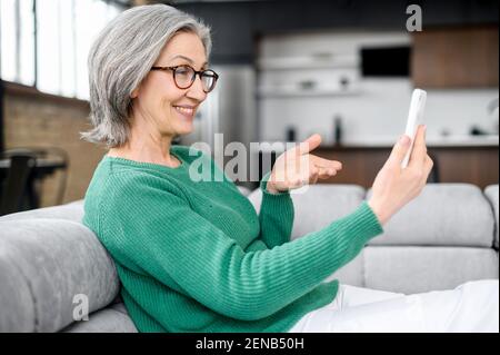 Moderne granny avec des rides magnifiques, regardant le téléphone, assis sur le canapé, parlant avec sa fille et voit son neveu, gesticultes, sur le salon, riant tout en regardant la vidéo drôle Banque D'Images