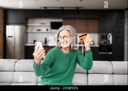 Moderne et ridules magnifiques, en utilisant le téléphone portable pour la vidéoconférence et en montrant sa nouvelle carte de crédit, des chiffons, fier d'elle-même, assis sur le canapé, en parlant avec la famille, dans le salon Banque D'Images