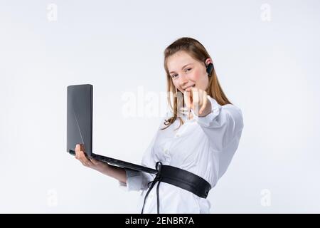 La jeune femme caucasienne attrayante se tient sur un fond gris isolé tient un ordinateur portable avec le sourire et les points de la main à la caméra Banque D'Images