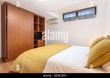 Chambre minimaliste, lit avec draps jaunes, face à une armoire en bois ouverte près d'un climatiseur. Banque D'Images