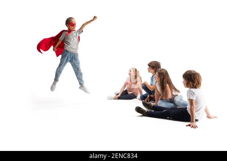 Puissance. Enfant prétendant être un super-héros avec ses amis assis autour d'elle. Les enfants sont excités, inspirés par leur ami robuste et courageux, sous un manteau rouge isolé sur fond blanc. Rêves, émotions concept. Banque D'Images