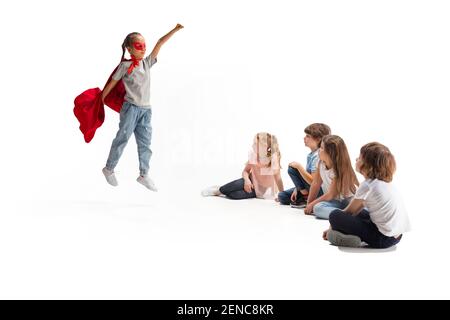 Puissance. Enfant prétendant être un super-héros avec ses amis assis autour d'elle. Les enfants sont excités, inspirés par leur ami robuste et courageux, sous un manteau rouge isolé sur fond blanc. Rêves, émotions concept. Banque D'Images