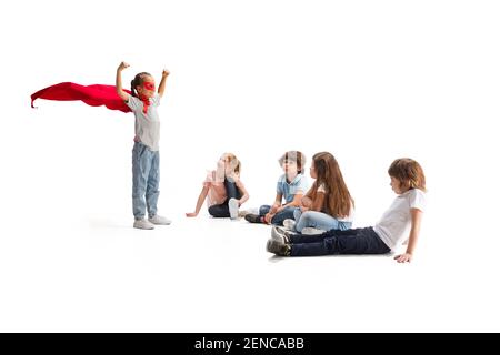 Puissance. Enfant prétendant être un super-héros avec ses amis assis autour d'elle. Les enfants sont excités, inspirés par leur ami robuste et courageux, sous un manteau rouge isolé sur fond blanc. Rêves, émotions concept. Banque D'Images