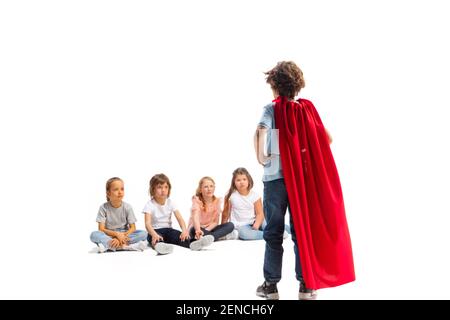 Puissance. Enfant prétendant être un super-héros avec ses amis assis autour de lui. Les enfants sont excités, inspirés par leur forte amie en robe rouge isolée sur fond blanc. Rêves, émotions concept. Banque D'Images