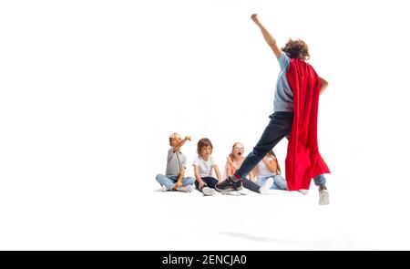 Puissance. Enfant prétendant être un super-héros avec ses amis assis autour de lui. Les enfants sont excités, inspirés par leur forte amie en robe rouge isolée sur fond blanc. Rêves, émotions concept. Banque D'Images