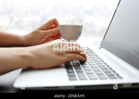 Une femme boit du café assis au clavier de l'ordinateur portable sur fond de fenêtre. Tasse de boisson chaude dans la main de la femme, pause pendant le travail Banque D'Images