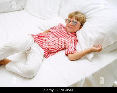 Portrait d'un bébé souriant dans une chemise à carreaux et avec des lunettes de soleil couchés sur un lit blanc. En vacances un jour d'été. Banque D'Images