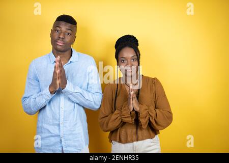 Jeune couple afro-américain debout sur fond jaune mendiant et priant avec les mains avec l'expression d'espoir sur le visage très émotionnel et Banque D'Images