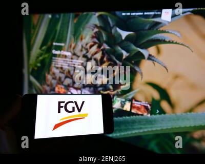 Personne tenant un téléphone portable avec le logo de la société d'agriculture malaisienne FGV Holdings Berhad à l'écran en face de la page Web. Mise au point sur l'affichage du téléphone. Banque D'Images