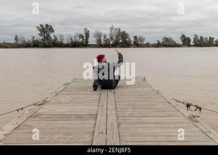 Un jeune homme de derrière porte une casquette rouge et un le chandail gris est assis dans un quai à côté d'un rivière prenant des selfies par jour nuageux Banque D'Images