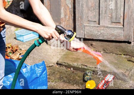 La mère lave les jouets des enfants à l'aide d'un tuyau d'eau, scène de style de vie à l'extérieur. Nettoyer les jouets des enfants avec de l'eau à l'extérieur, en été, par temps ensoleillé, en gros plan. Banque D'Images