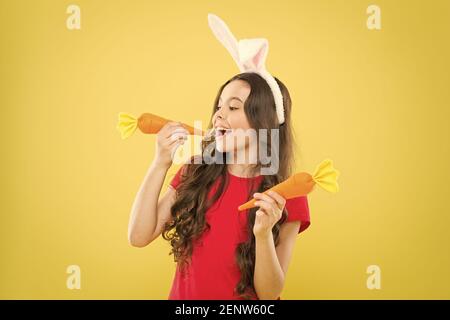 petite fille heureuse nourriture pour lapins. enfance en bonne santé. bonne pour vos dents. Grignotez une carotte comme un lièvre. Les enfants dans les oreilles de lapin aiment manger de la carotte. Costume de lapin enfant avec carotte. Banque D'Images