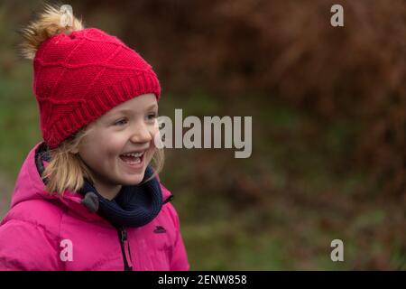 Jeune fille en manteau d'hiver rose et chapeau rouge marchant dans la forêt, riant, fond vert, promenade d'hiver dans les bois, lachendes Mädchen im Wald, Hope Banque D'Images