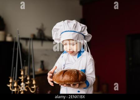 Concept de saine alimentation. Un petit garçon heureux cuisine dans la cuisine par une belle journée d'été. Lors d'un pique-nique, le boulanger mange du pain et des bagels dans un tablier blanc Banque D'Images