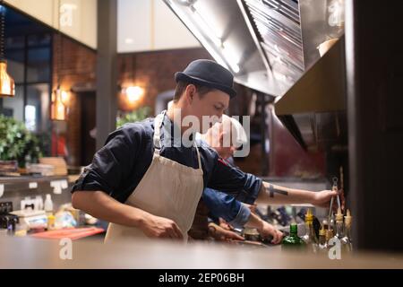 Jeune homme en chapeau et en tablier prenant des pinces pour cuisiner plat près des collègues dans la cuisine de la cafétéria Banque D'Images