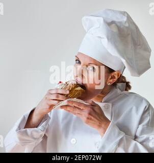 Une femme chef goûtant une pâtisserie dans un manteau et un chapeau de chef blanc traditionnel. Banque D'Images