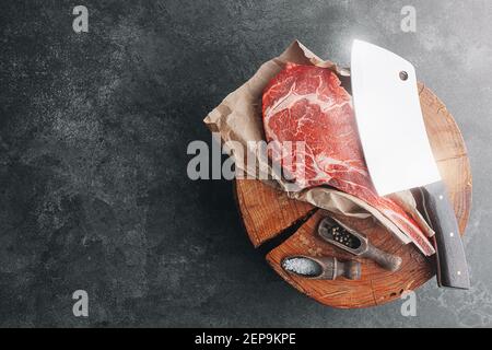 sur un bloc de bois pour la viande un steak de tamahawk frais cru ou un steak de cowboy avec une hache de boucher pour la viande, à côté de lui est un mélange de poivrons et Banque D'Images