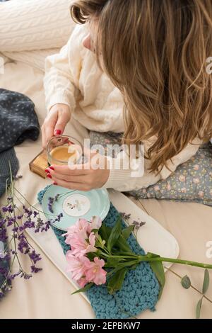 Femme habillée de laine, elle tient une tasse de café avec les mains pendant qu'elle est posée sur le lit. Banque D'Images