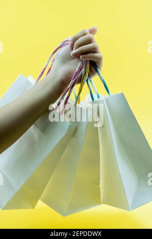 Photo courte de la main d'un enfant tenant un bouquet de différents sacs de shopping vierges colorés sur fond jaune isolé. Concept de solde du Vendredi fou. Copier sp Banque D'Images