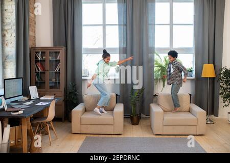Deux mignons frères et sœurs de race mixte en vêtements décontractés sautant et dansant fauteuils confortables et grandes fenêtres dans la salle de séjour tout en s'amusant Banque D'Images