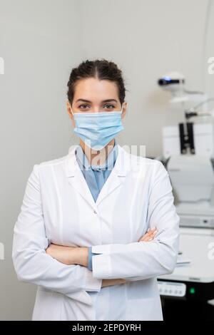 Jeune ophtalmologiste réussi dans le masque protecteur et blancoat la croisant les bras près de la poitrine tout en se tenant contre l'équipement médical Banque D'Images