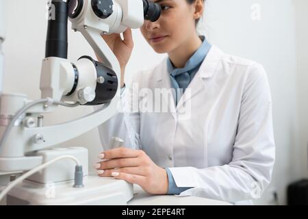 Jeune femme ophtalmologiste en whitecavoine utilisant de nouveaux équipements médicaux à vérifiez la vue de l'un des clients dans les cliniques contemporaines Banque D'Images