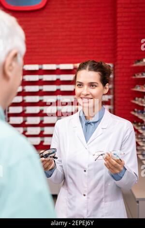 Jeune femme souriante ophtalmologiste en blancoat tenant deux paires de de nouvelles lunettes tout en aidant l'homme âgé avec le choix des lunettes Banque D'Images