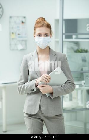 Jeune gestionnaire de bureau élégant en costume et tenue de masque de protection tablette par poitrine tout en se tenant face à l'appareil photo contre son lieu de travail Banque D'Images