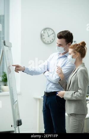 Jeune gestionnaire confiant dans le masque de protection pointant vers le tableau blanc pendant expliquer les données financières à son collègue de highligter Banque D'Images