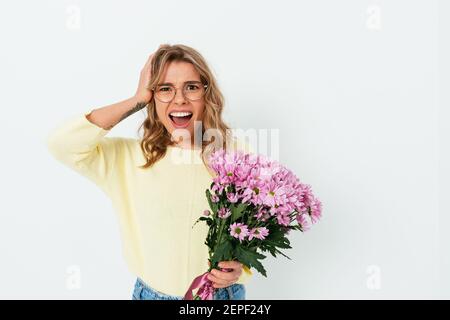 Jeune femme malheureuse en lunettes et vêtements décontractés portant un bouquet de fleurs et de la tête avec l'expression du visage perplexe sur bac studio blanc Banque D'Images