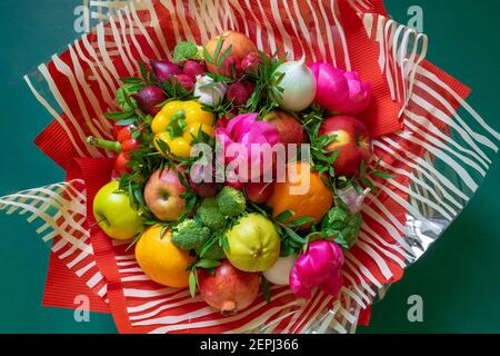 Un bouquet de légumes et de fruits aux couleurs vives et multicolores avec du rose pivoines et feuilles vertes enveloppées dans du papier rouge ondulé et cellophane clair à rayures Banque D'Images