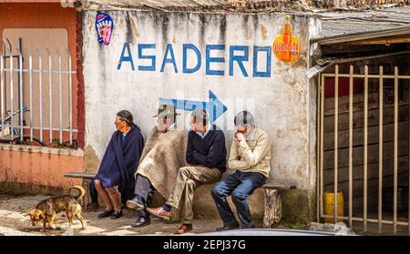 1 chien, colombien 3 hommes, 1 femme assis sur le banc, en attente. 1 dans le poncho traditionnel à chapeau, Ruana. Asadero signe Boyacá département, Colombie, Amérique du Sud Banque D'Images